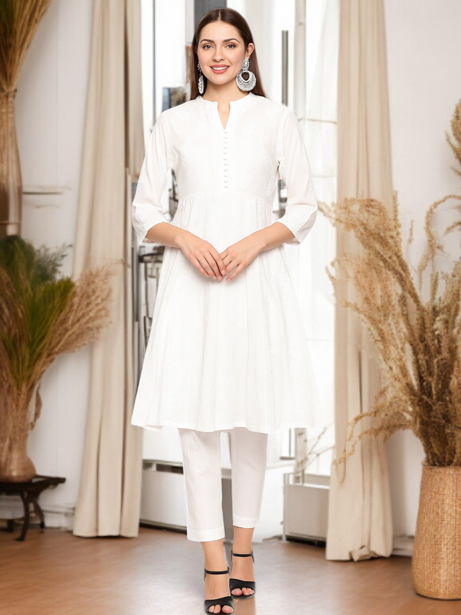 Woman in a white outfit standing in a room with decorative plants and curtains.