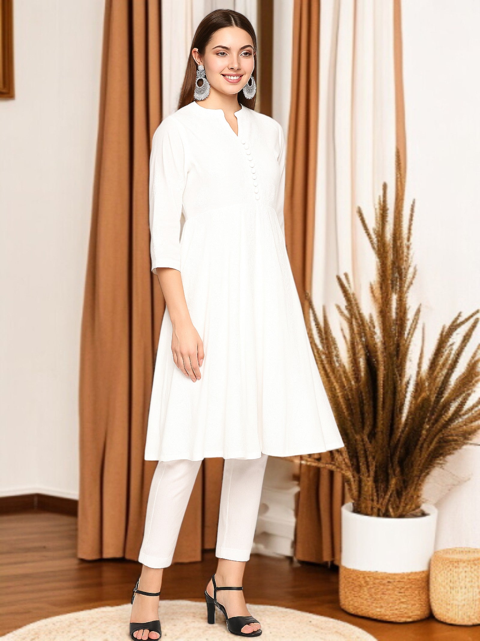 Woman wearing a white outfit standing in a room with brown curtains and decorative plants.